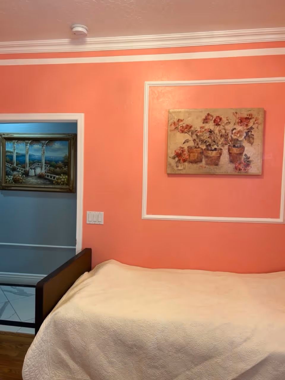 A bedroom with coral-colored walls featuring white trim. There is a bed with a white quilted blanket in the foreground. On the wall above the bed is a painting of potted flowers. Through an open doorway, another room with a framed painting of a scenic outdoor view is visible.