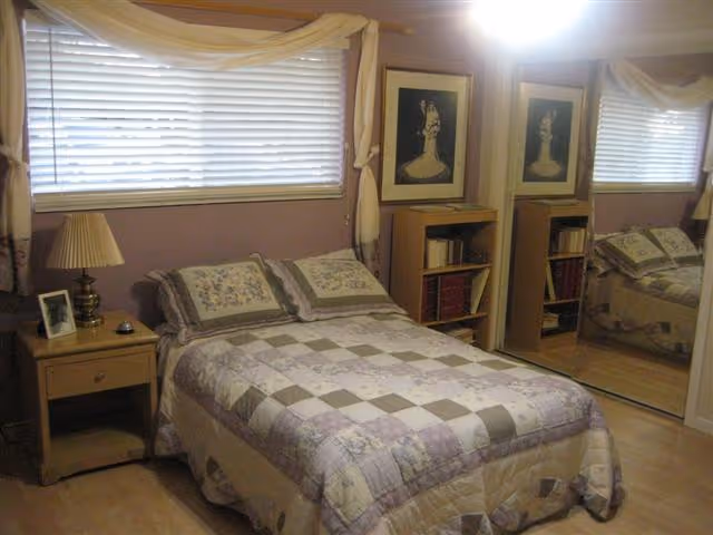 A cozy bedroom with a double bed covered in a patchwork quilt in shades of beige, lavender, and gray. There are two matching pillows on the bed. To the left of the bed is a wooden nightstand with a lamp, a framed photo, and a small object. Above the bed is a window with closed blinds and light-colored curtains. On the right side of the room, there is a mirrored closet door reflecting the bed and part of the room. Two framed artworks hang on the wall next to the closet.