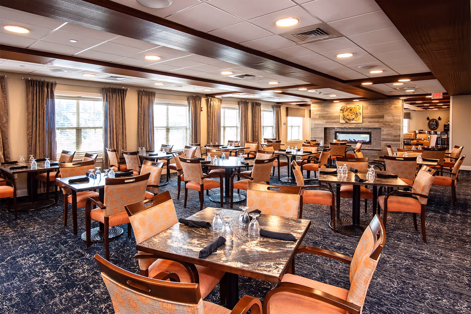 Spacious, well-lit dining room with multiple set tables and upholstered chairs, large windows, and a fireplace.