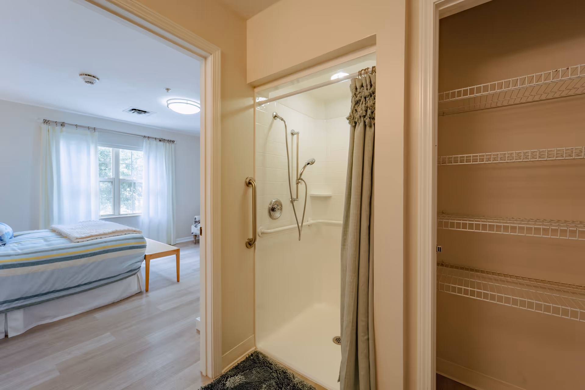 A bright bedroom with a bed by a window, an adjacent open shower with grab bars, and a nearby closet with wire shelving.