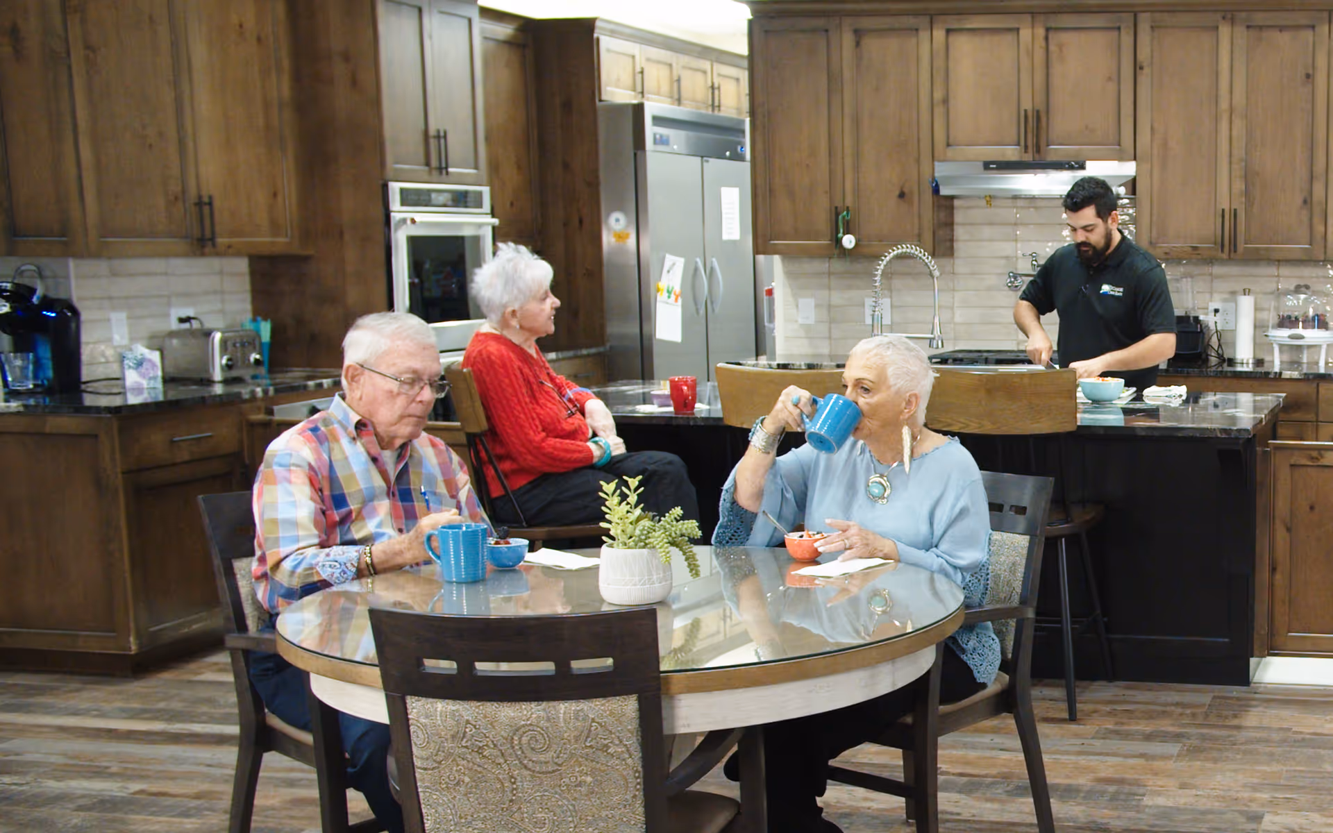 Three elderly individuals sitting around a round glass-top dining table in a kitchen area. One woman in a light blue top is drinking from a blue mug, an elderly man in a plaid shirt is eating from a bowl, and another woman in a red sweater is sitting with her arms crossed. In the background, a man in a black shirt is preparing food at the kitchen counter with wooden cabinets and stainless steel appliances.