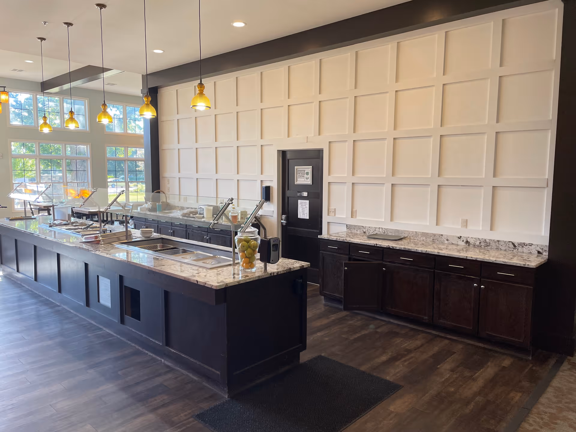 Interior view of a modern dining buffet area with a long marble countertop island featuring food serving stations with glass sneeze guards. The room has large windows letting in natural light, pendant lights hanging from the ceiling, dark wood cabinetry, and a decorative white paneled wall with a black door.
