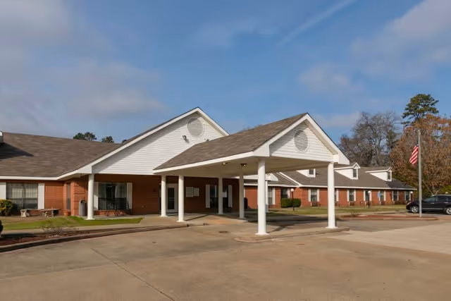 Exterior view of a single-story healthcare and rehabilitation center building with a covered entrance supported by white columns, a paved driveway, and an American flag on a flagpole to the right. The building has a brick lower facade and white siding above, with a clear blue sky overhead.