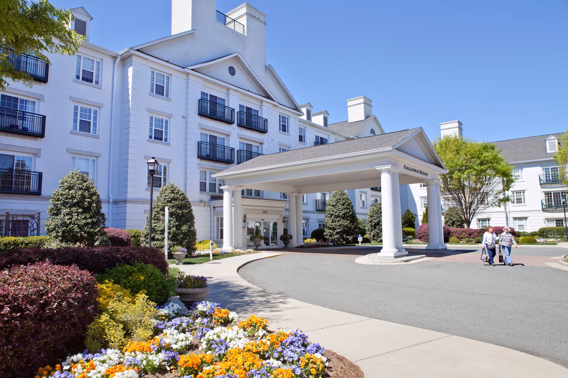 Exterior view of Galloway Ridge at Fearrington, a large white multi-story building with balconies and a covered entrance. There are colorful flower beds and neatly trimmed bushes in the foreground, and two people walking on the driveway near the entrance under a clear blue sky.