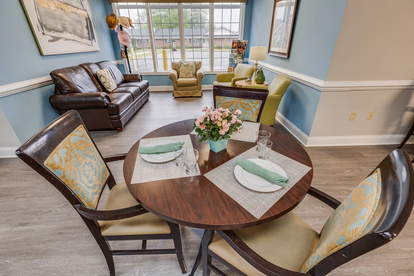 Round wooden dining table set with plates and napkins in a bright assisted-living common room with chairs, a leather sofa and armchairs by a large window.