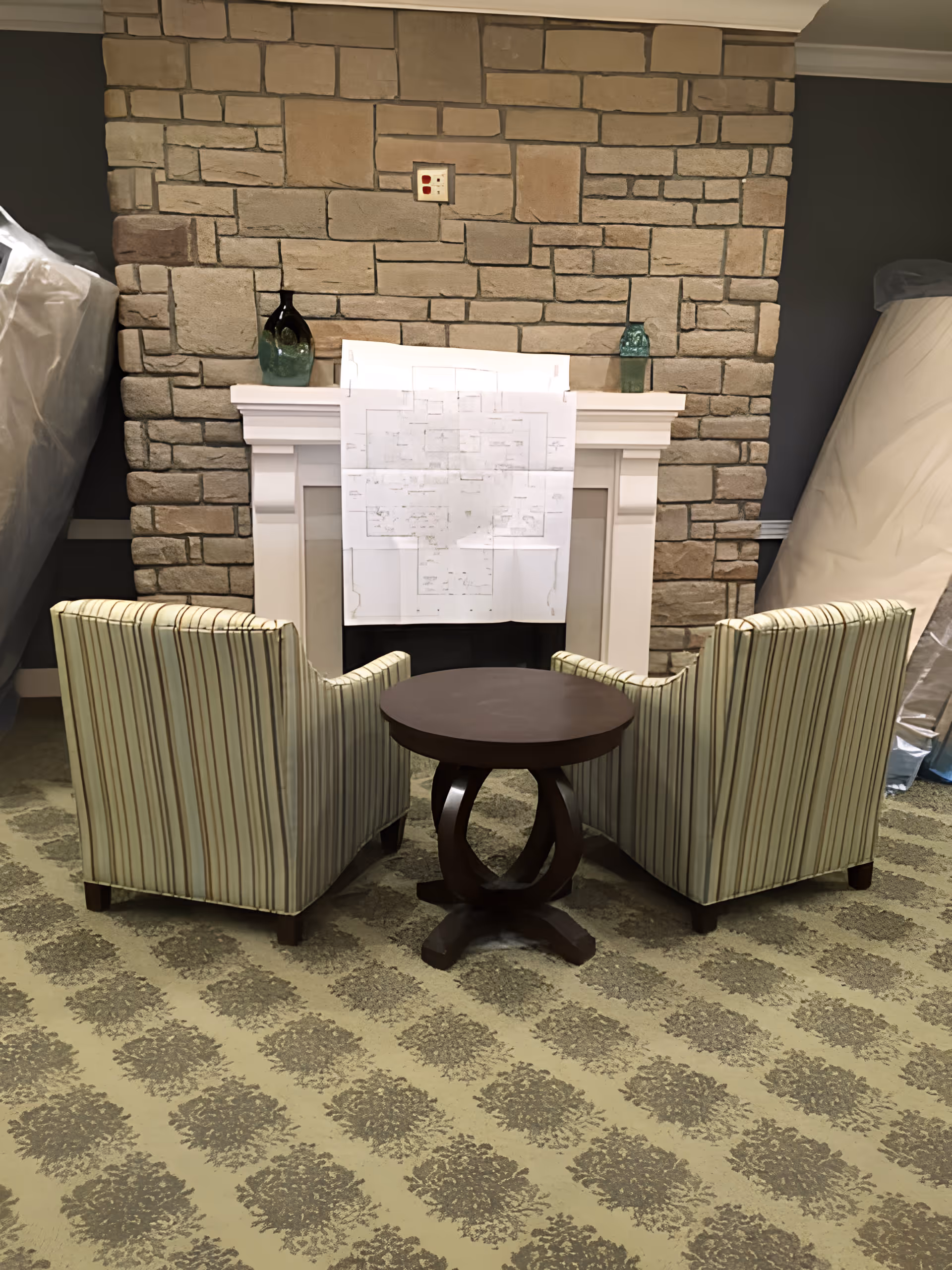 Two striped armchairs facing a small round wooden table in front of a white fireplace with a stone wall background. A floor plan is taped to the fireplace mantel, and two decorative vases are placed on either side of the mantel. The room has patterned carpet and mattresses covered in plastic are visible on both sides.