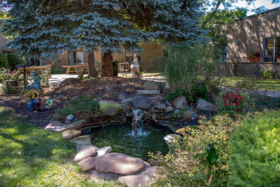 A peaceful outdoor garden area at Sodus Rehabilitation and Nursing Center featuring a small pond with a water fountain surrounded by rocks and greenery. There are trees, bushes, flowers, and garden decorations around the pond. In the background, there are picnic tables and a brick building.