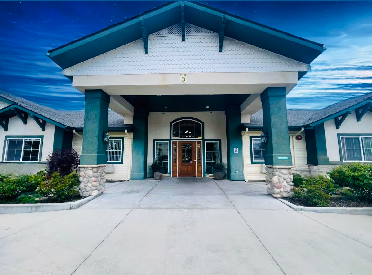 Front entrance of a building with a covered porch supported by green pillars with stone bases, wooden double doors with glass panels, and surrounding windows. The building has a pitched roof and landscaped greenery on either side of the entrance.