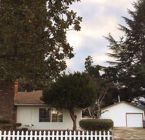 A single-story residential building with a white picket fence in front, a large tree partially obscuring the view, and a detached garage with a closed door. The sky is partly cloudy and there are other trees around the property.