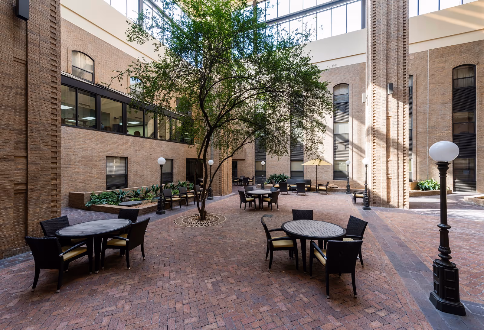 A spacious indoor courtyard area with brick flooring and brick walls, featuring several round tables with black wicker chairs arranged around them. There is a tree growing in the center with a metal grate around its base, and several black lamp posts with white globes providing lighting. Large windows and tall columns are visible along the walls, and natural light fills the space from above.