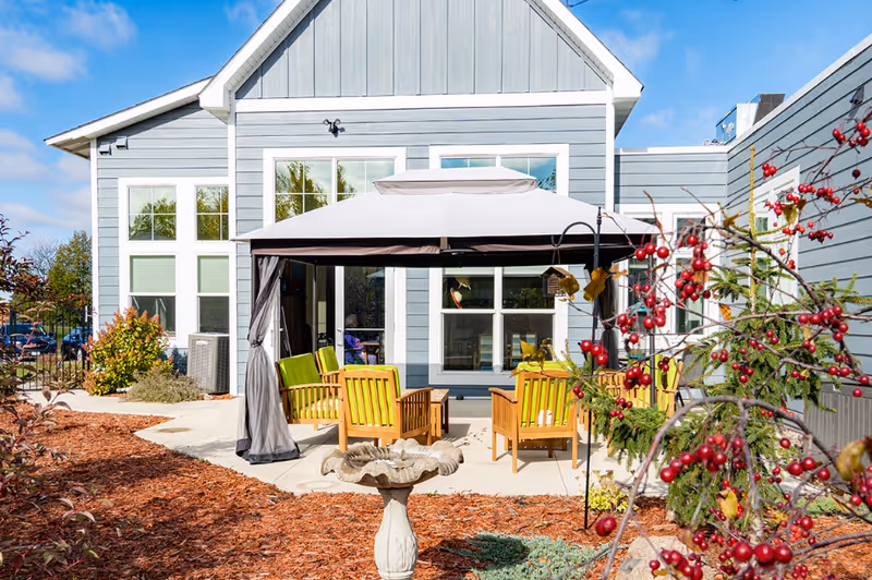 Outdoor patio area at a senior living facility with wooden chairs featuring green cushions arranged under a large canopy. The patio is surrounded by landscaping with red berries on bushes and a birdbath in the foreground. The building exterior is light blue with white trim and large windows.