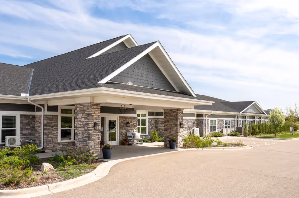 Exterior view of a senior living facility named Rivers of Life featuring a covered entrance with stone pillars, large windows, and a paved driveway with parking spaces. The building has a dark shingled roof and is surrounded by landscaped greenery under a partly cloudy sky.