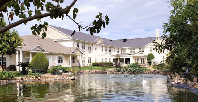 Two-story assisted living building set behind a landscaped pond with a fountain and a white swan.