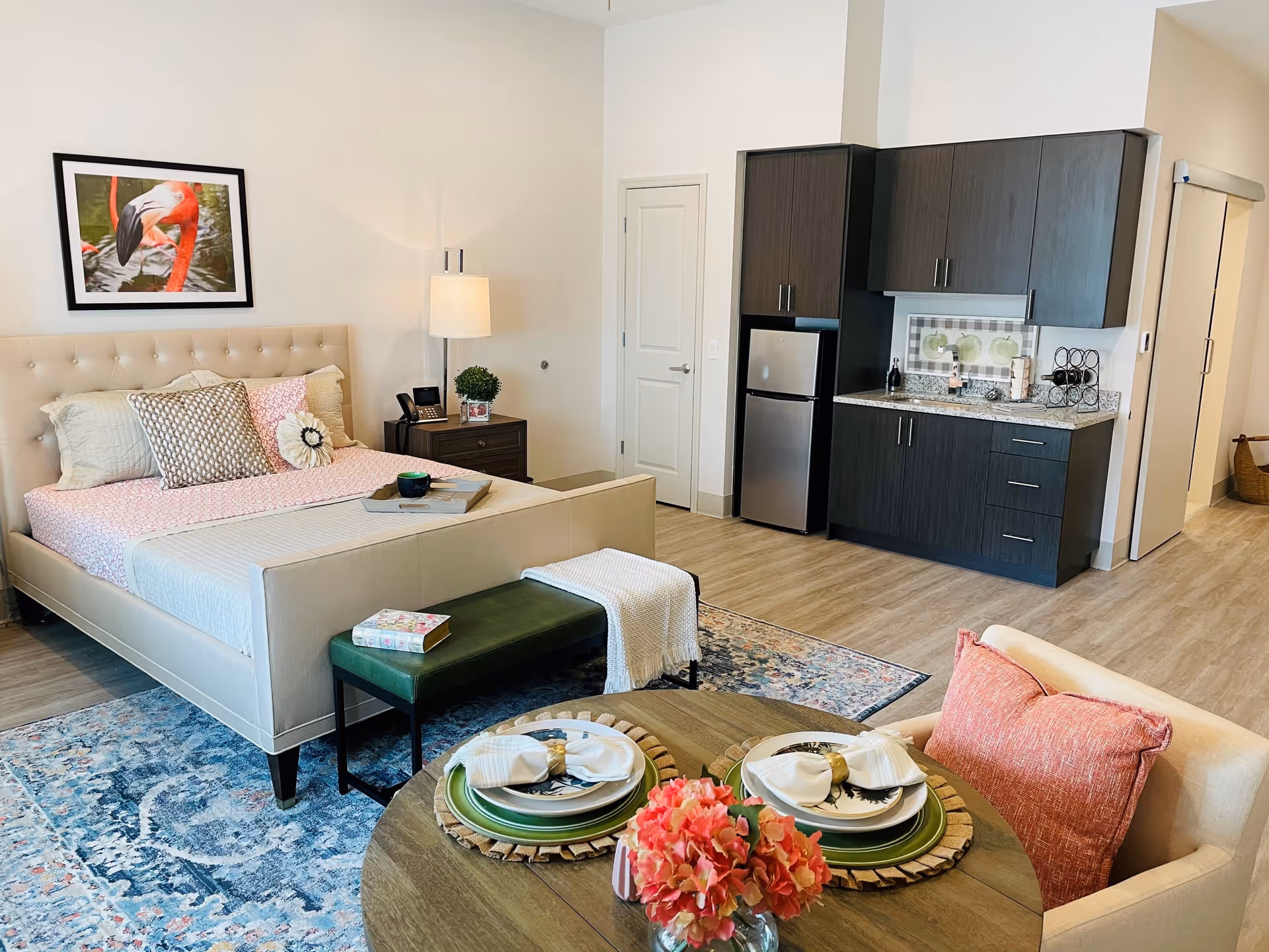 A studio apartment interior featuring a beige upholstered bed with multiple pillows and a pink bedspread, a dark green bench at the foot of the bed, a wooden nightstand with a lamp and phone, a small kitchenette with dark cabinets and a stainless steel mini fridge, and a round dining table set for two with plates, napkins, and a pink flower centerpiece. The room has light wood flooring and a colorful area rug under the bed.