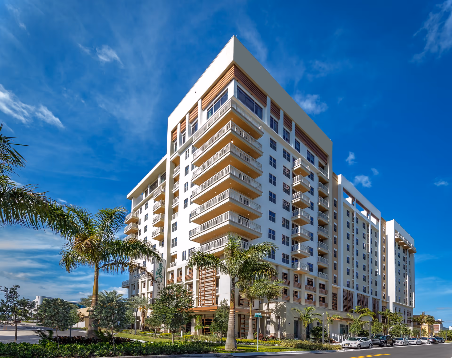 Exterior view of a multi-story modern senior living facility building with balconies, palm trees, and a clear blue sky in Fort Lauderdale.
