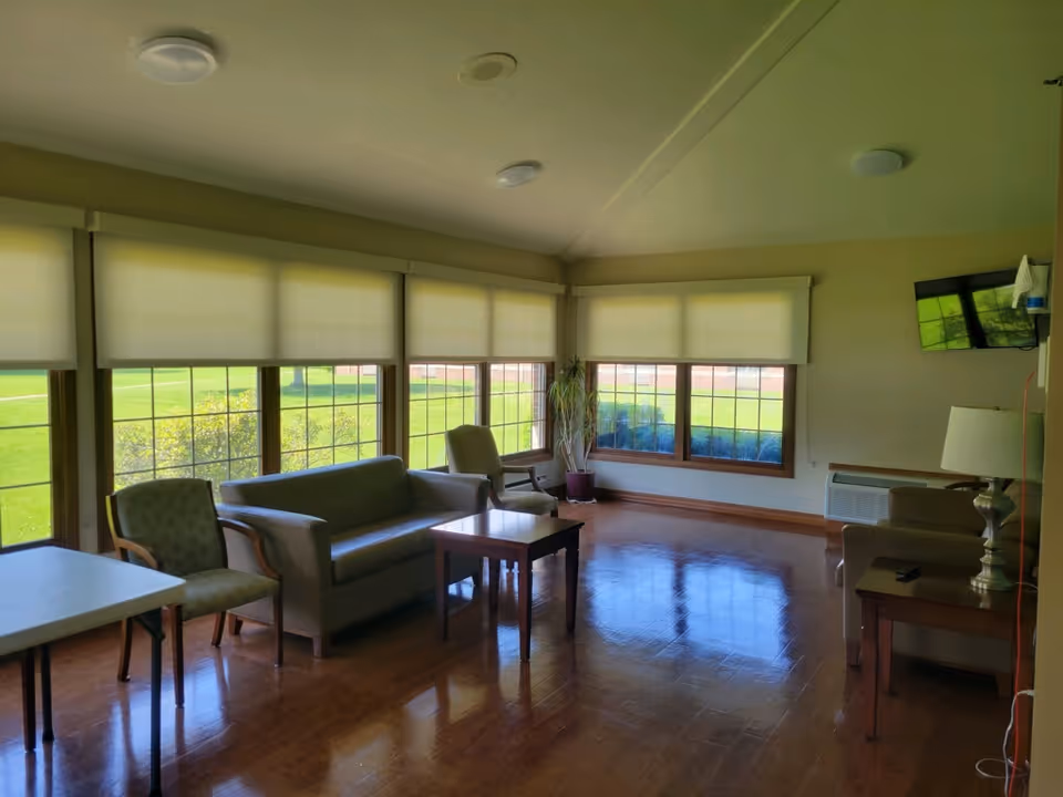A bright living room area with large windows covered by light-colored roller shades, allowing natural light to fill the space. The room contains a gray sofa, two armchairs, a wooden coffee table, a side table with a lamp, a wall-mounted TV, and a potted plant in the corner. The floor is wooden and polished, reflecting the light from the windows.