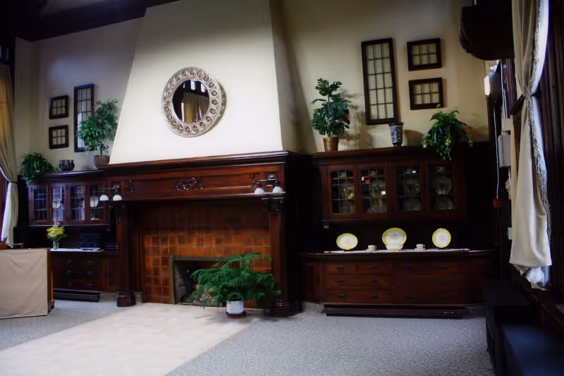 Common room with a large dark-wood fireplace mantel between glass-front cabinets, decorative plates, potted plants, and framed mirrors.