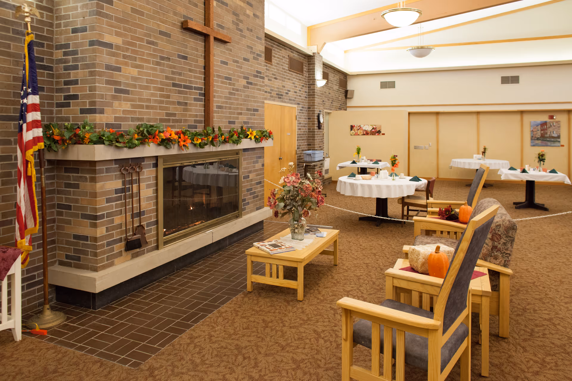 Interior of a senior living facility common area with a large brick fireplace decorated with a garland of autumn leaves and flowers. There are wooden chairs with cushions and small pumpkins on the side tables, a coffee table with a vase of flowers and magazines, and round tables covered with white tablecloths and small floral centerpieces in the background. An American flag stands to the left of the fireplace.