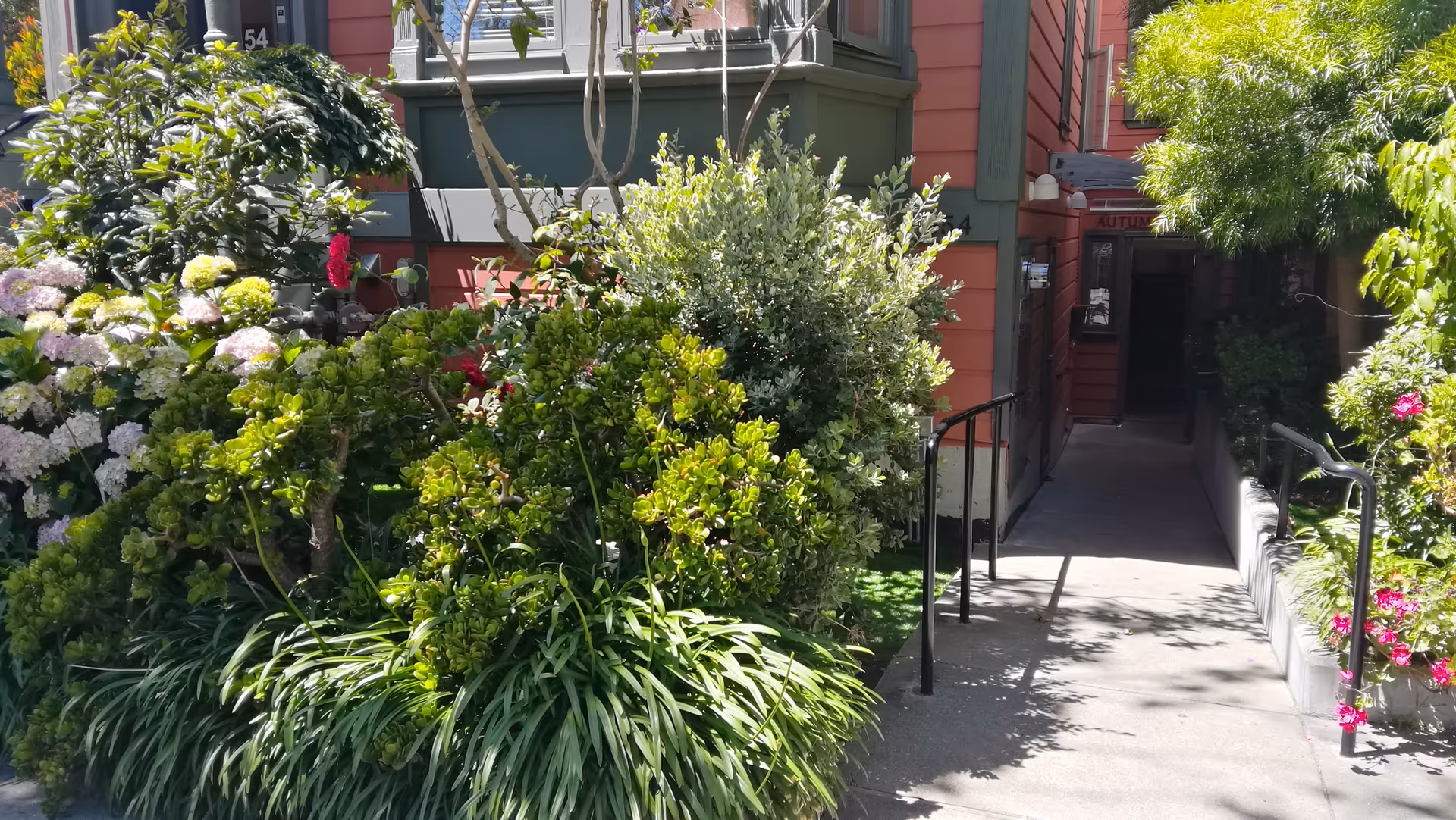 A garden pathway leading to the entrance of a building with red exterior walls. The pathway is flanked by lush green bushes and flowering plants, with handrails on both sides. The entrance door is partially visible in the background.