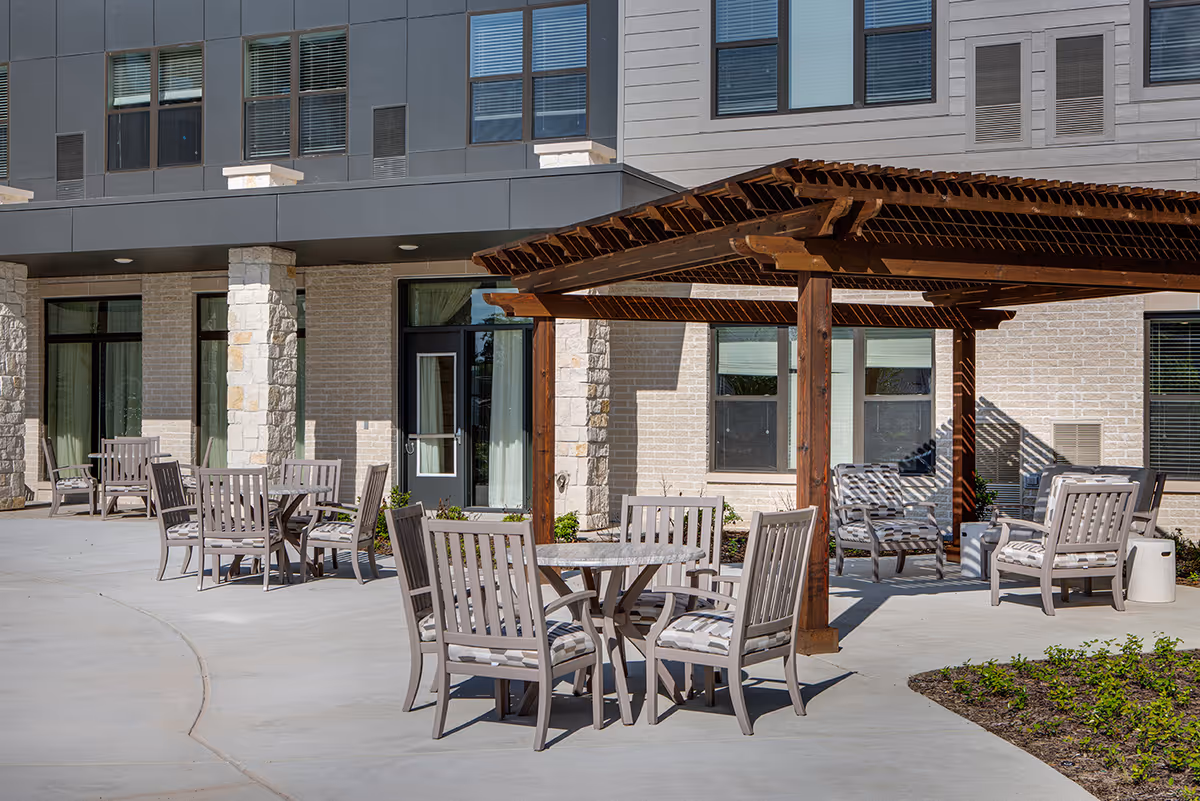 Outdoor patio area at Fountainwood at Lake Houston featuring multiple tables and chairs with cushions, a wooden pergola providing partial shade, and the exterior wall of the building with windows and a door.