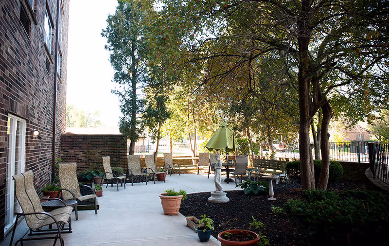Outdoor patio area at Autumn Ridge Rehabilitation Centre with multiple cushioned chairs arranged along a brick building and around a concrete patio. There are several potted plants, a small statue, a green umbrella, and trees providing shade in the space.