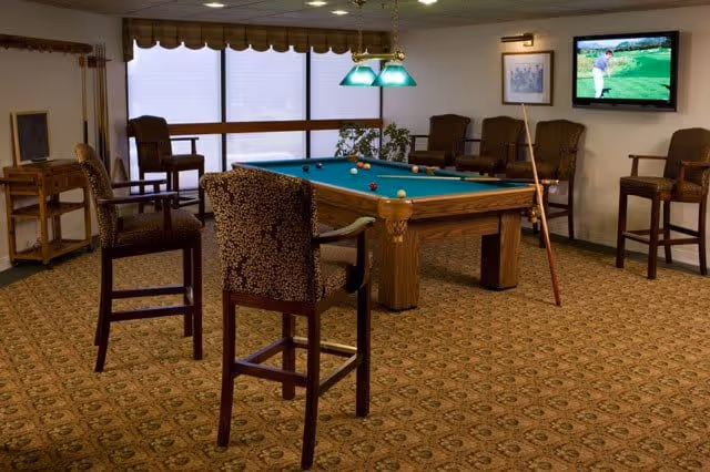 A cozy indoor recreation room featuring a pool table with billiard balls and cues, surrounded by several upholstered chairs and bar stools. A flat-screen TV mounted on the wall displays a golf game. The room has patterned carpet flooring, large windows with blinds, and framed artwork on the walls.