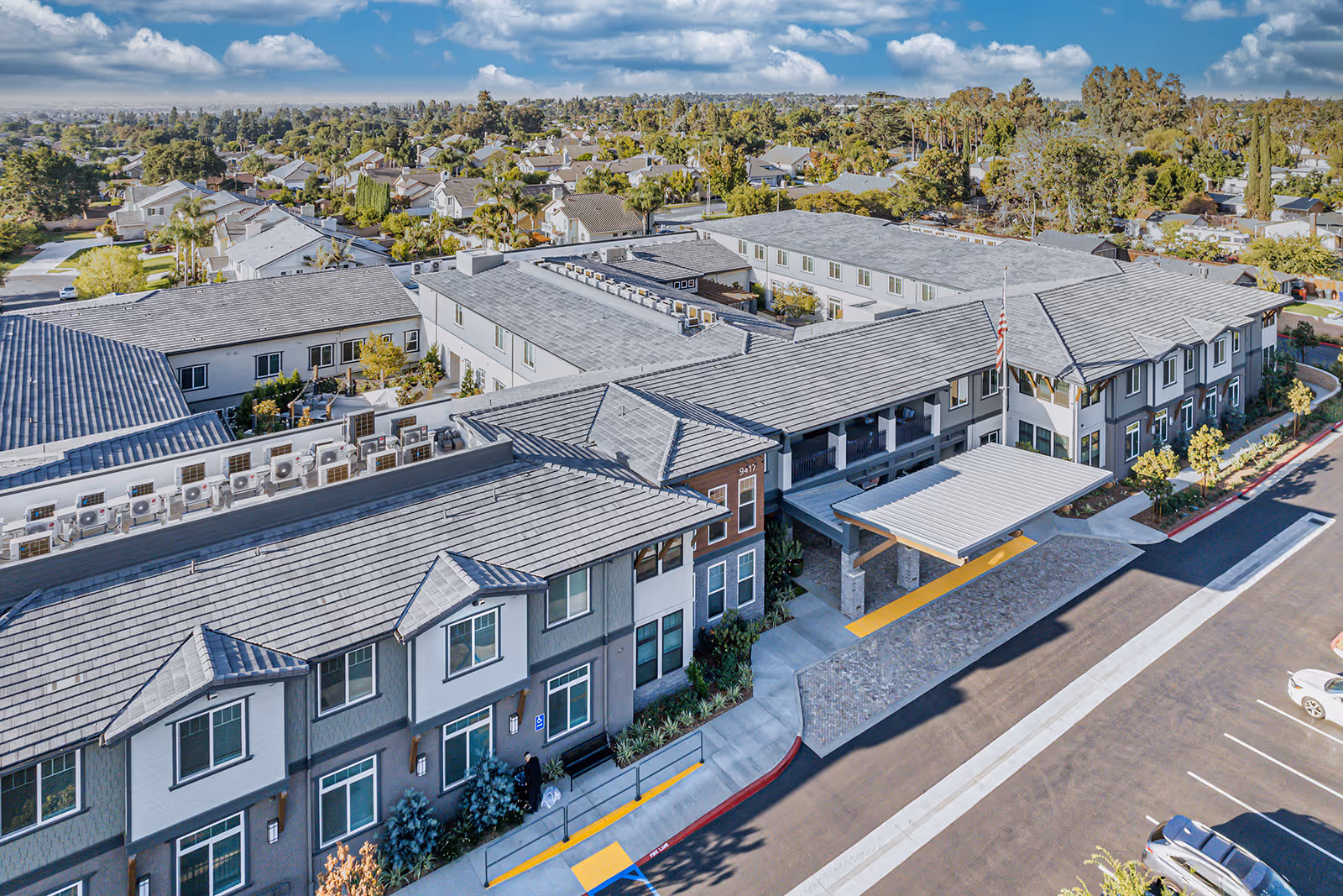 Aerial view of Allara Senior Living facility showing a large, modern two-story building with gray roofing and multiple windows. The entrance has a covered drop-off area with a flagpole flying the American flag. Surrounding the building are landscaped areas and a parking lot with several cars. In the background, a residential neighborhood with many houses and trees is visible under a partly cloudy sky.