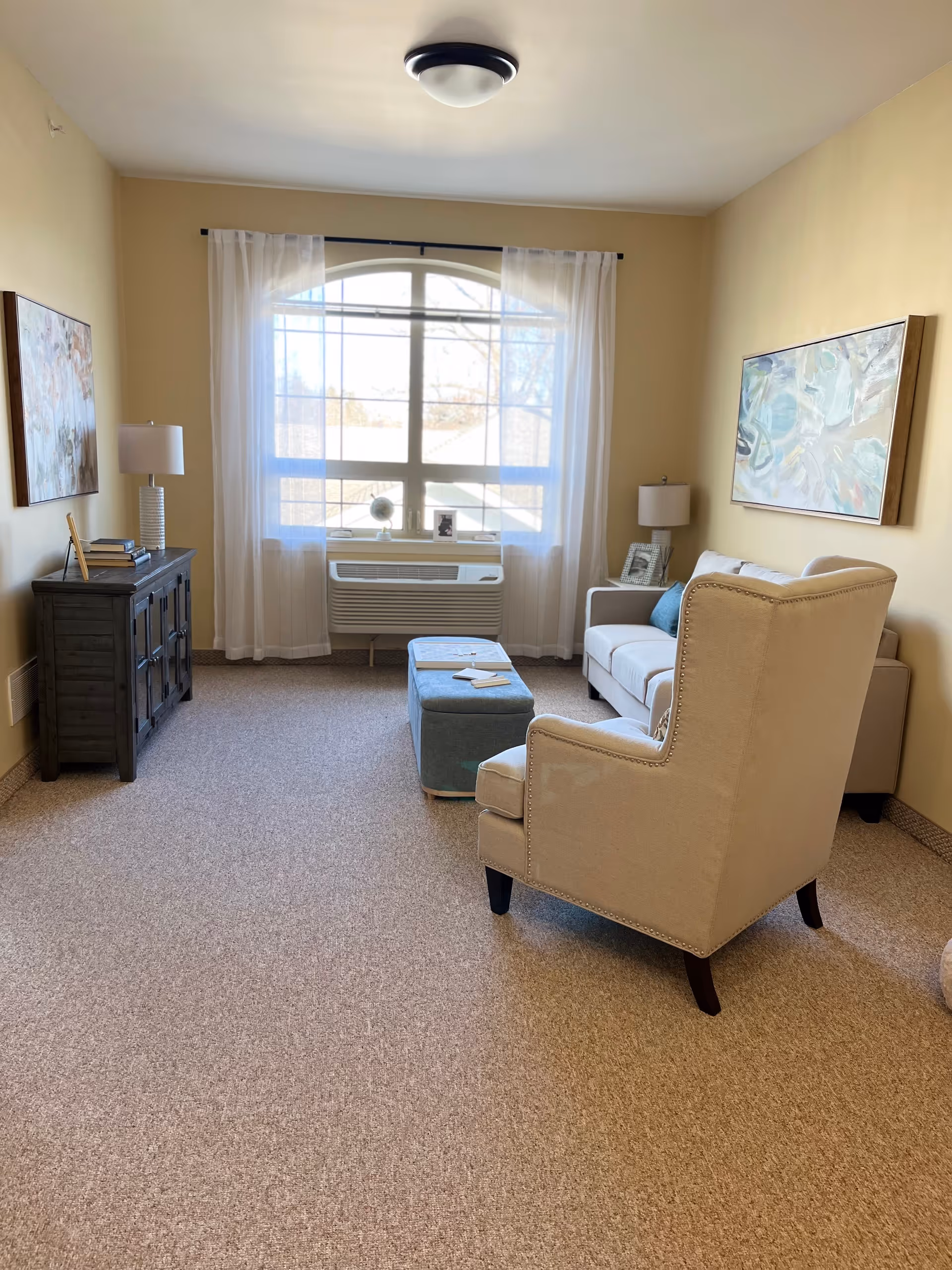 A cozy living room with beige walls and carpeted floor. The room features a large window with sheer white curtains, a white armchair, a white sofa with blue cushions, a blue ottoman, a dark wooden cabinet with books and a lamp on top, and framed artwork on the walls.