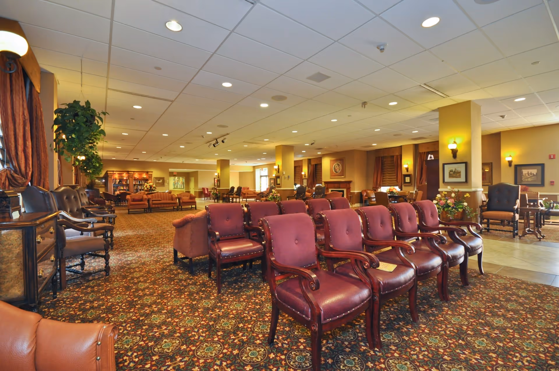 Spacious furnished common lounge with rows of burgundy upholstered chairs, sofas, and decorative patterned carpeting.