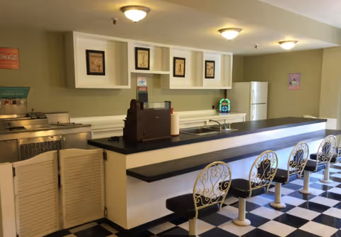 Retro-style communal dining counter with five decorative swivel stools, checkerboard floor, and a kitchen area behind the counter.