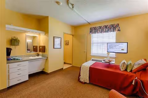 Yellow-walled bedroom in a senior living facility with a red bed, kitchenette, and TV.