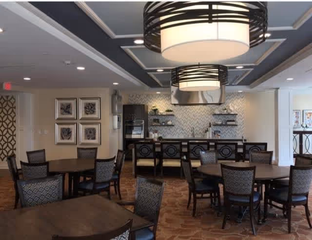 A spacious dining room with multiple round wooden tables and cushioned chairs arranged neatly. The room features large modern pendant lights hanging from the ceiling, a patterned carpet, and a wall with decorative framed artwork. In the background, there is a kitchen area with a tiled backsplash, open shelves, and a stainless steel range hood.