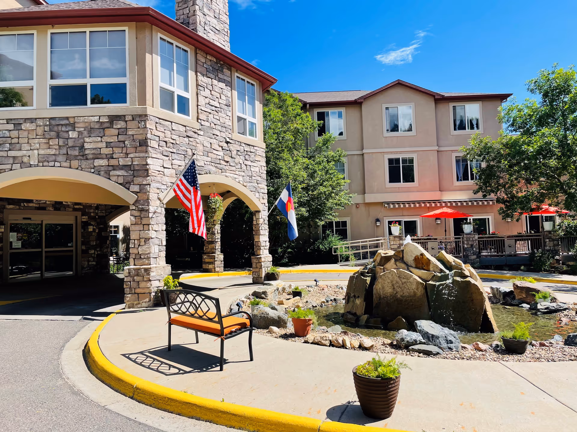 Exterior view of Elk Run Assisted Living facility showing a stone and stucco building with large windows, an entrance with arches, two flags (American and Colorado), a small pond with rocks and a fountain, a bench with an orange cushion, potted plants, and red umbrellas on a patio area under a clear blue sky.