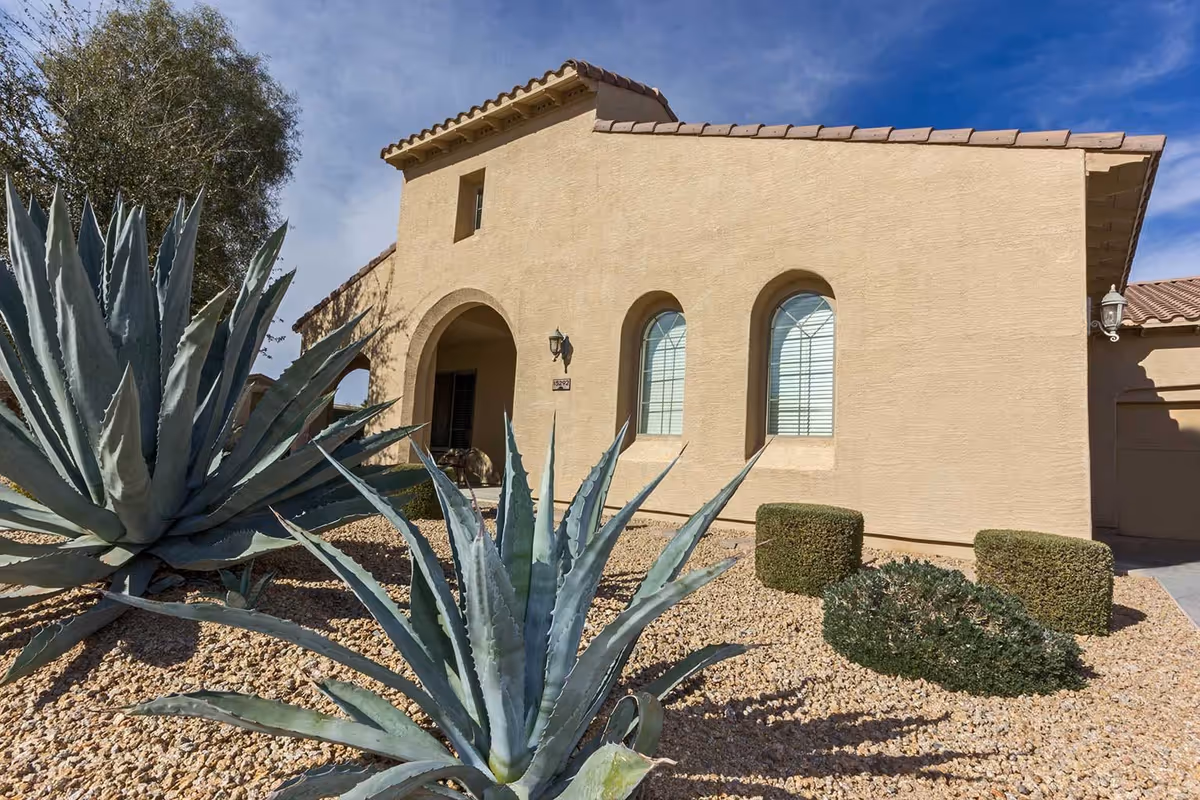 Exterior view of a beige stucco building with a tiled roof, featuring arched windows with blinds and a covered porch area. The foreground shows desert landscaping with large agave plants, small bushes, and gravel ground cover under a blue sky with some clouds.