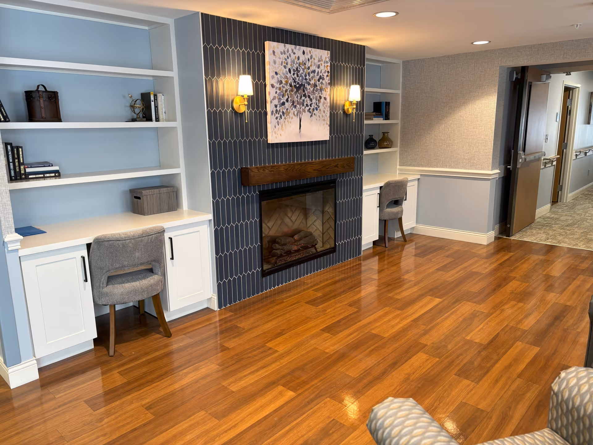 A cozy interior space with polished wooden floors, featuring a fireplace with a dark tiled surround and a painting of a tree above it. On either side of the fireplace are built-in white shelves and desks with gray upholstered chairs. The walls are painted in soft blue and beige tones, and there is a hallway visible in the background.