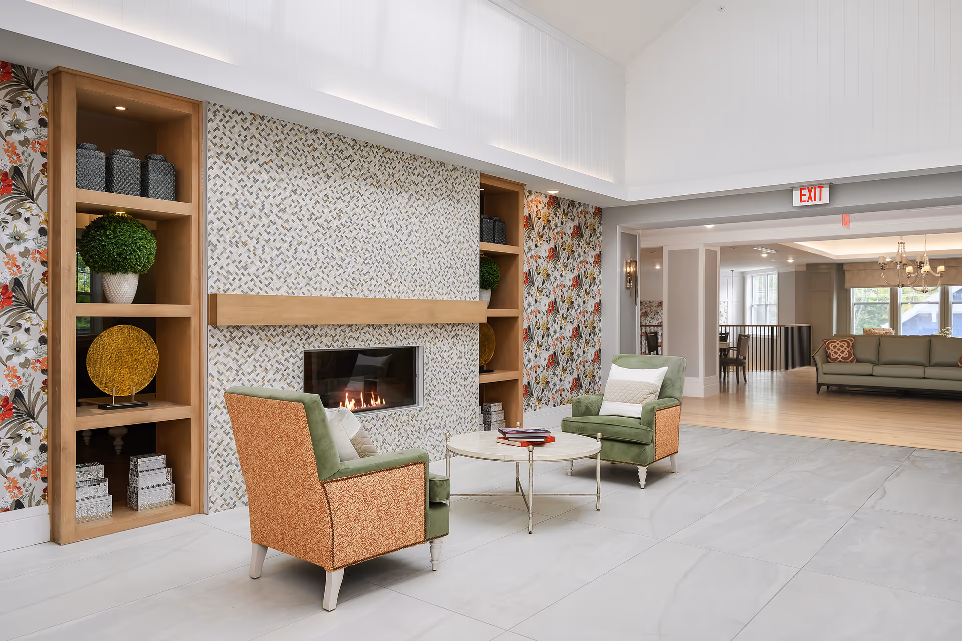 A bright and spacious living room area with two upholstered armchairs facing a modern fireplace set in a tiled wall. The fireplace is flanked by built-in wooden shelves decorated with plants, boxes, and decorative items. The room features floral wallpaper on the side walls, large light-colored floor tiles, and an open view into another seating area with a sofa and chandelier. An exit sign is visible above a doorway.