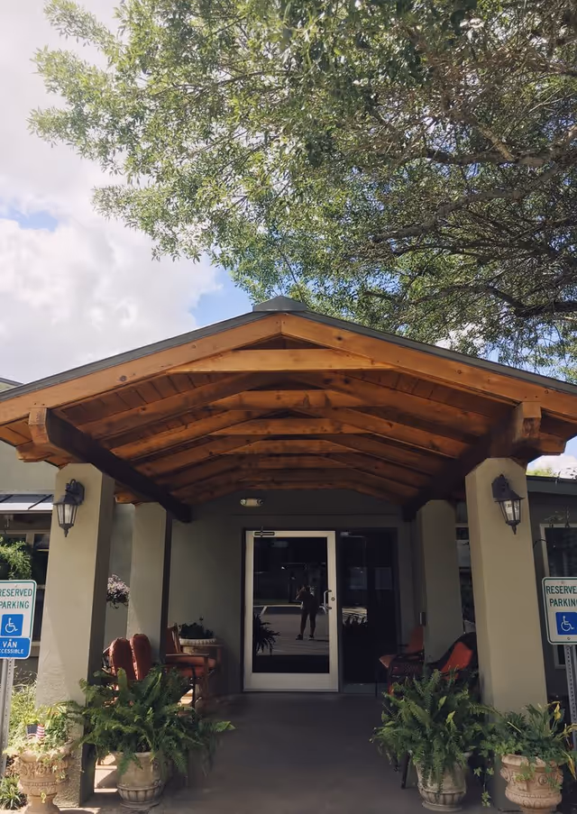 Entrance to a building with a wooden peaked roof supported by columns, two reserved parking signs for handicapped parking on either side, potted plants and chairs near the entrance, and a large tree overhead.