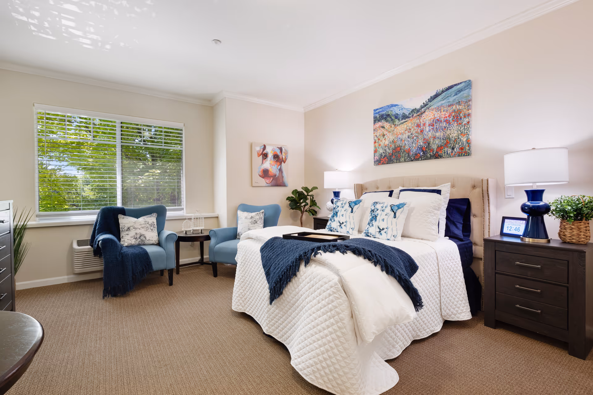 Bright furnished bedroom with a made bed, two blue armchairs by a window, bedside tables and wall artwork.