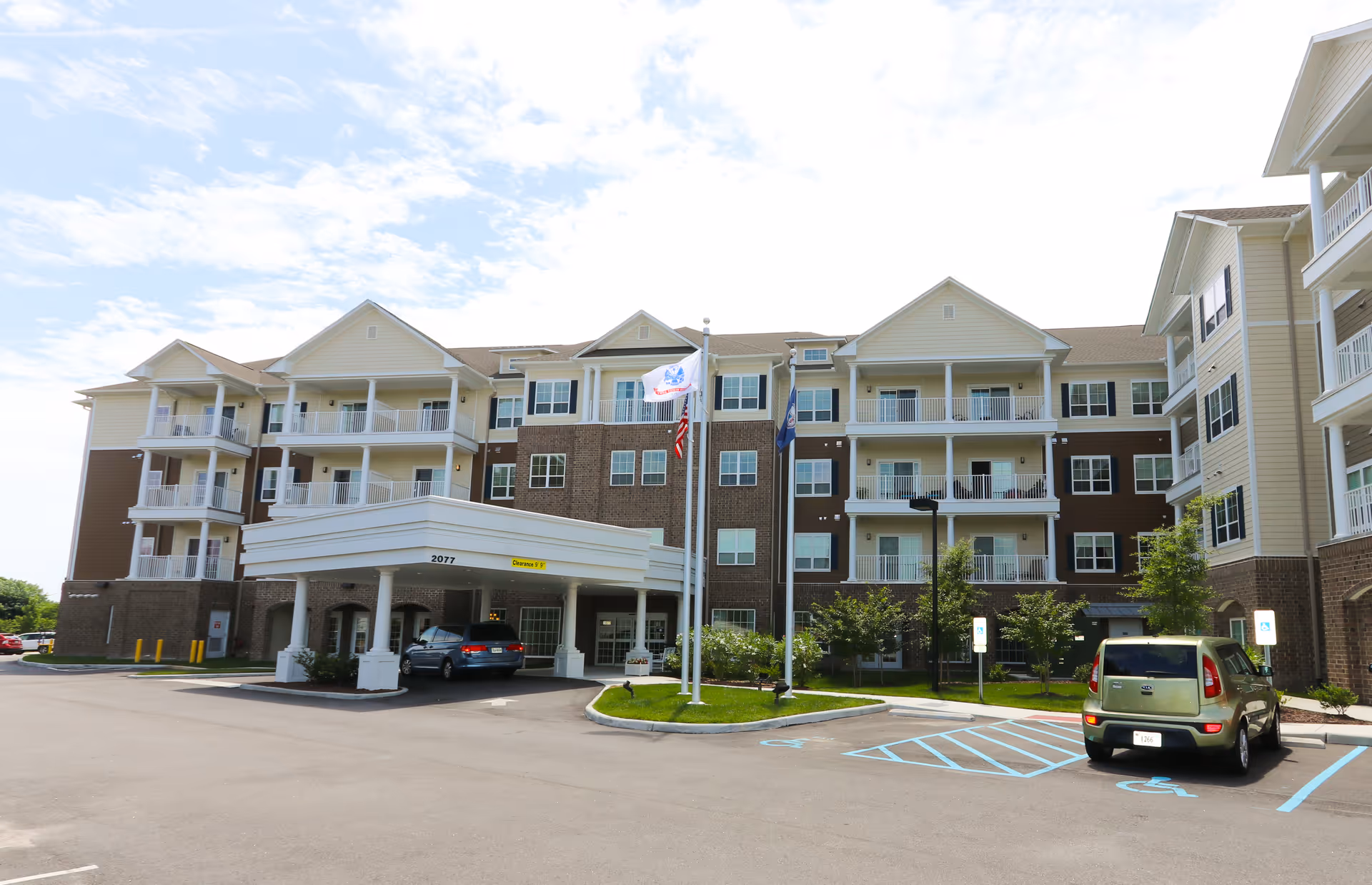 Exterior view of a multi-story senior living facility named Harmony at Independence with balconies, a covered entrance, three flagpoles, and a parking lot with handicap parking spaces.