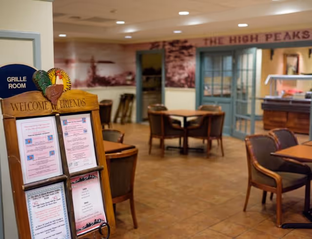 Interior view of a dining area with several tables and chairs arranged on a tiled floor. A wooden stand near the entrance displays menus and a sign that says 'WELCOME FRIENDS' with a decorative turkey on top. A blue sign on the wall indicates the 'GRILLE ROOM'. In the background, there are double glass doors and a wall mural with the words 'THE HIGH PEAKS'.