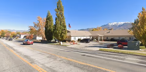 Exterior view of a single-story assisted living facility with a beige roof and walls, an American flag in front, several parked cars, and trees with autumn foliage. Snow-capped mountains are visible in the background under a clear blue sky.