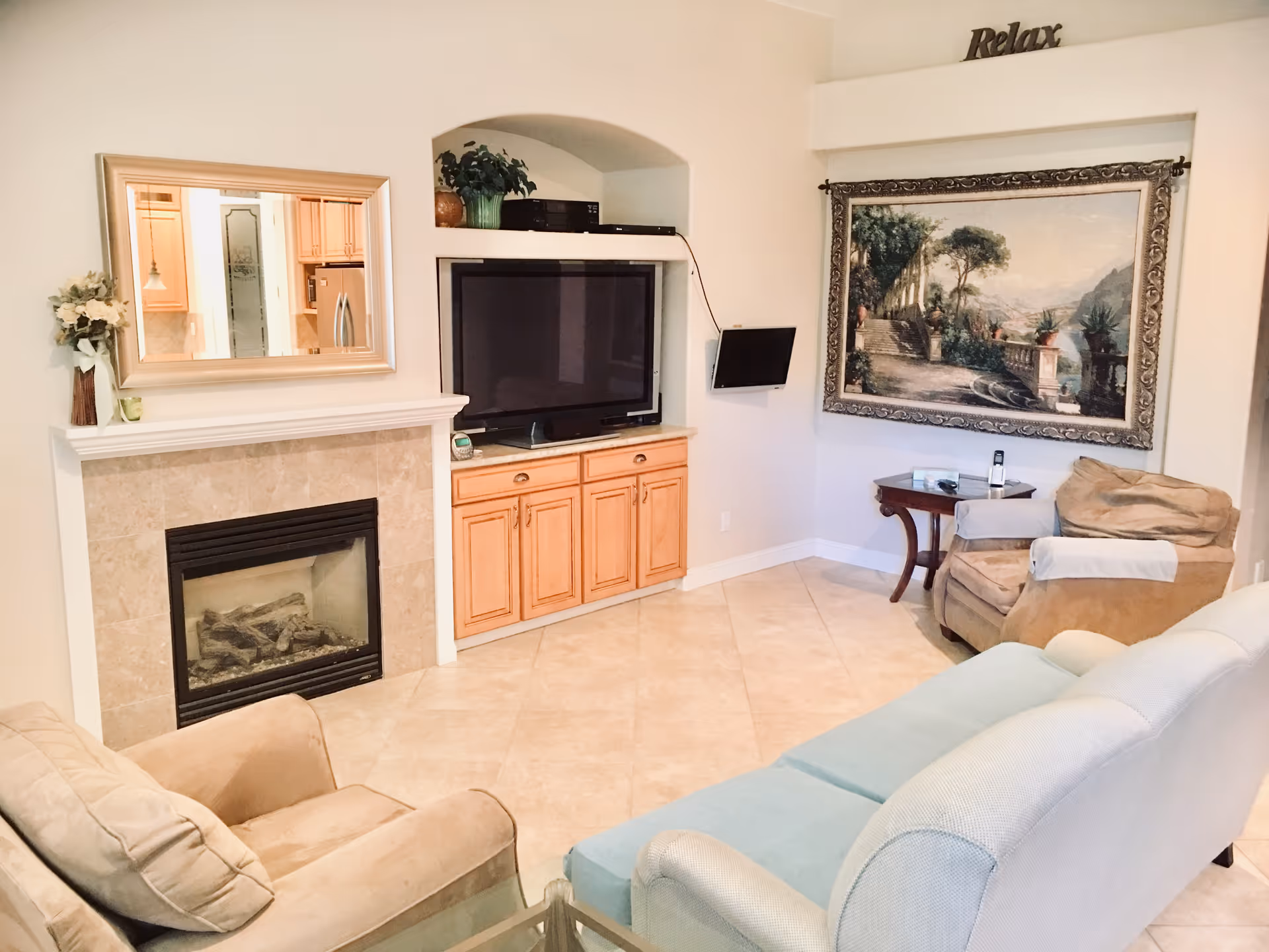A cozy living room with beige tiled flooring, a fireplace with a beige tile surround, a large flat-screen TV on a wooden cabinet, a beige armchair, a light blue sofa, a small wooden side table, a large framed painting of a scenic outdoor terrace, and a decorative 'Relax' sign on the wall above the painting.