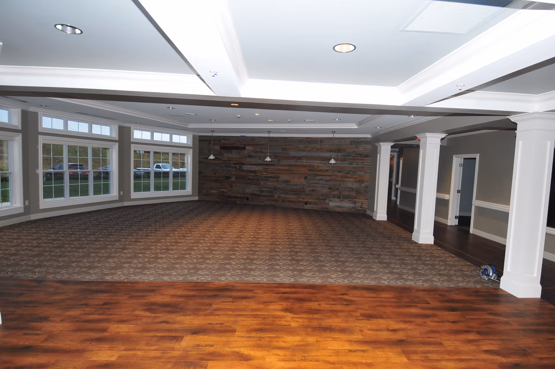 A spacious empty room with large windows on one side, a wooden accent wall at the far end, carpeted flooring in the main area, and wood flooring in the foreground. The ceiling has recessed lighting and white beams, with three pendant lights hanging near the wooden wall. White columns separate the carpeted area from a hallway with dark wood flooring.