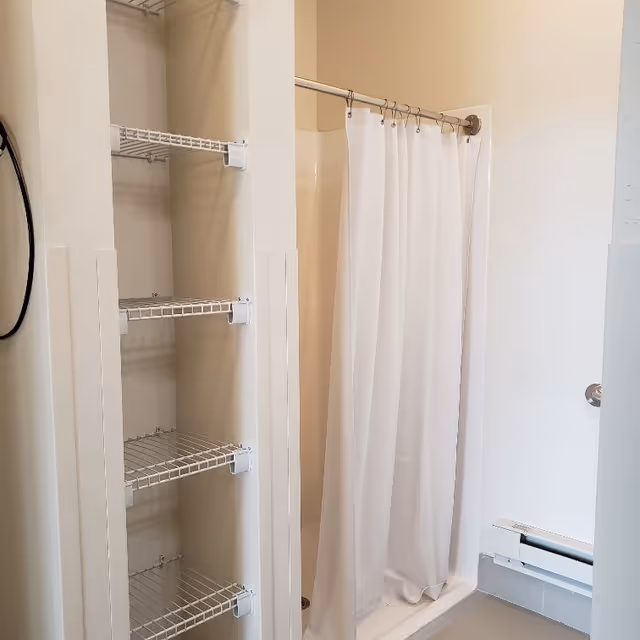 A bathroom with a white shower curtain hanging on a curved shower rod, a built-in shelving unit with wire shelves, and a white wall-mounted heater below a towel ring.