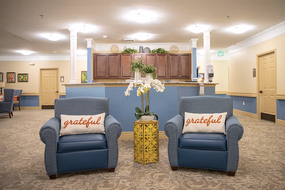 Two blue armchairs with 'grateful' pillows flanking a decorative plant in a senior living facility lobby with a reception desk behind.