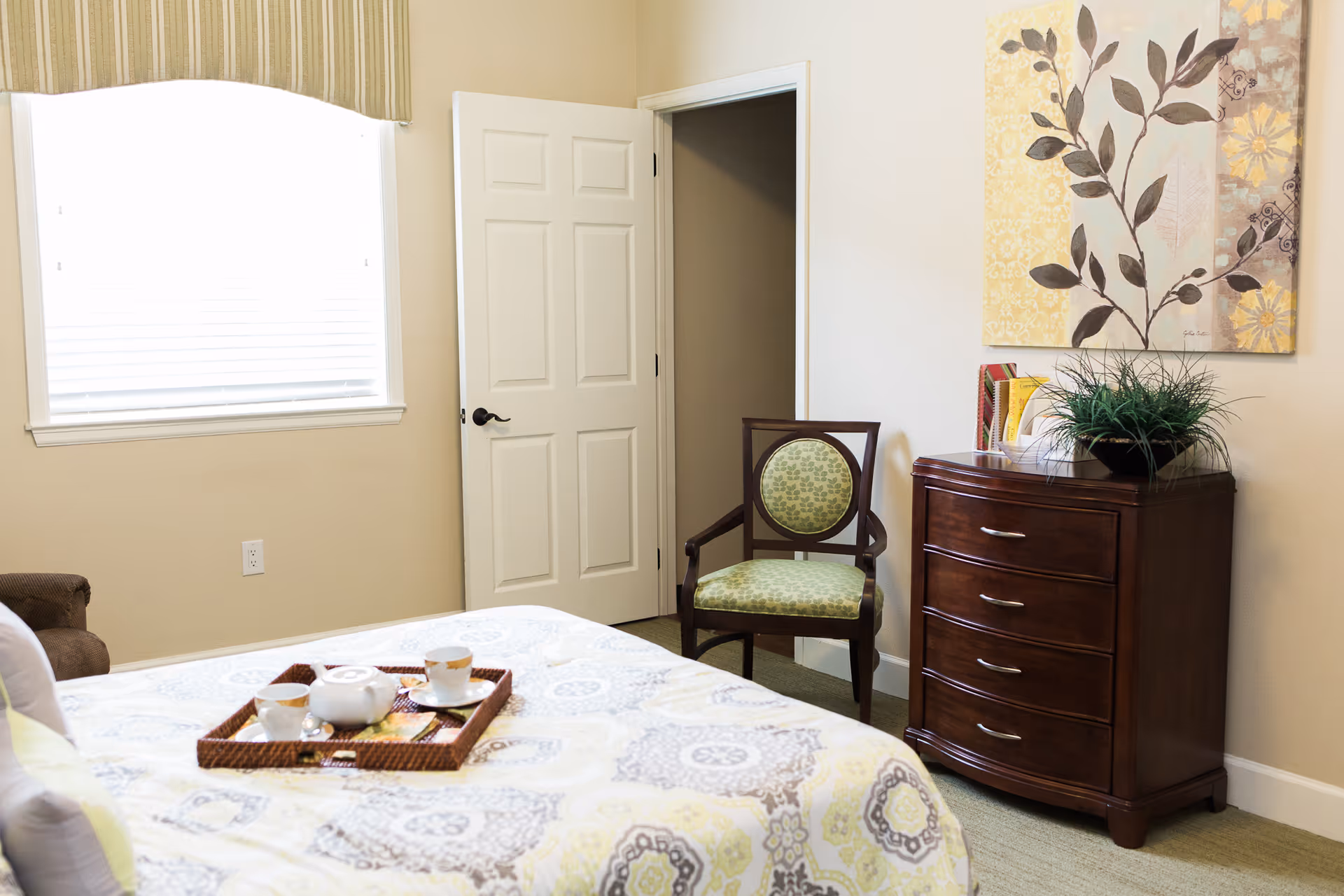 A cozy bedroom with a bed covered in a patterned quilt, a tray with a teapot and cups on the bed, a wooden chair with green patterned upholstery, a dark wooden dresser with a plant and books on top, and a decorative floral painting on the wall. A window with blinds and a striped valance is on the left, and a white door is open in the background.