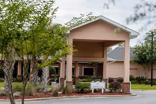 Entrance of Partin Place Senior Living facility showing a covered drop-off area with brick pillars, surrounded by landscaped greenery and trees under a cloudy sky.