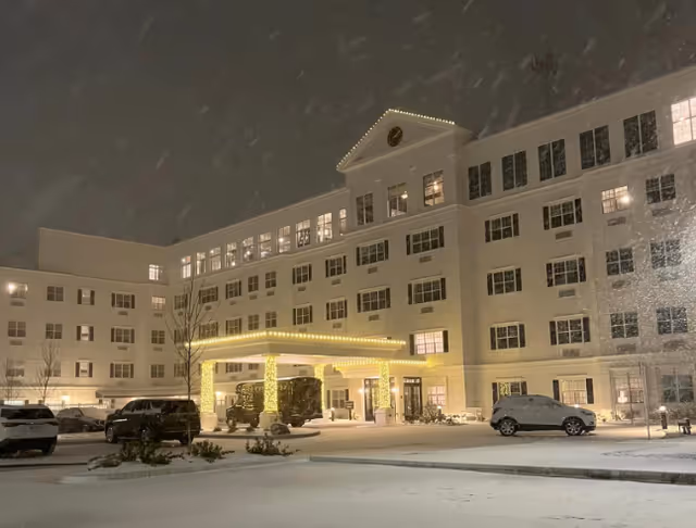Exterior front of a multi-story senior living building at night with an illuminated covered entrance, parked cars, and falling snow.