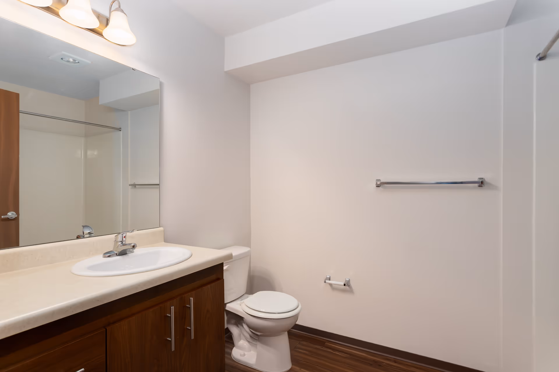 A clean bathroom with a white sink set in a beige countertop, wooden cabinets below, a large mirror above the sink, a toilet next to the sink, a towel rack on the wall, and a shower area with a curtain rod visible.