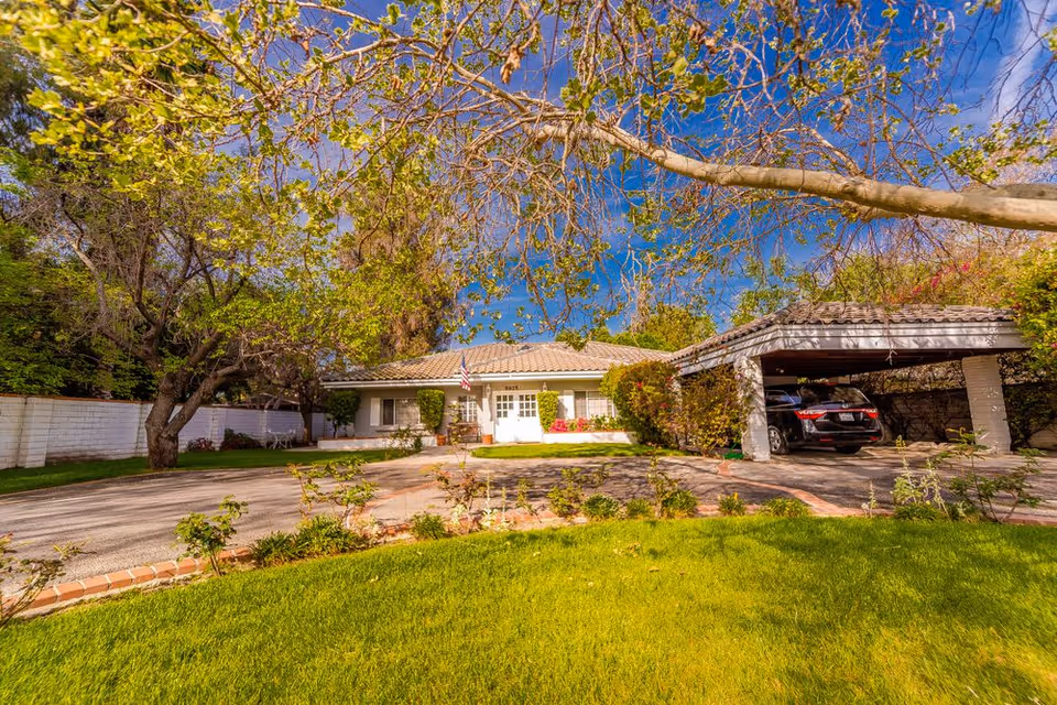 Front exterior view of a single-story residential building with a tiled roof, surrounded by trees and greenery. There is a driveway leading to a covered carport with a black car parked underneath. The lawn in the foreground is well-maintained with small plants along the edge of the driveway. The sky is clear and blue.